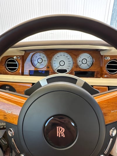 The image shows a close-up, interior view of a Rolls-Royce steering wheel and dashboard. The steering wheel is dark brown leather with a black center hub featuring the Rolls-Royce logo in a rose gold color. The dashboard is adorned with rich wood grain trim, chrome accents, and circular gauges. Visible gauges include a speedometer reading up to 260 km/h, a "Power Reserve %" gauge, and a fuel gauge. There are also circular air vents and various chrome buttons and controls. The environment appears to be well-lit, possibly from natural light, given the reflections on the surfaces. The scene is a static shot, focusing on the luxurious details of the car's interior. There is no visible indication of the city of Dub, Montenegro.