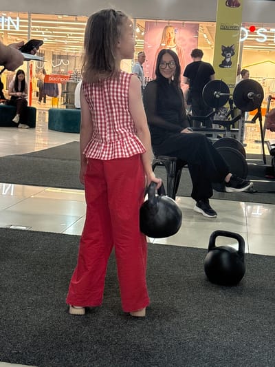 In Tri Protoka, Russia, a young girl stands facing away from the camera, wearing a red and white checkered peplum top and red pants. She holds a black kettlebell in her right hand. To her right, a woman with glasses and dark clothing sits on a chair next to weightlifting equipment, smiling at the camera. Another black kettlebell rests on the carpeted floor in front of them. The background suggests an indoor location, possibly a shopping mall or gym, with storefronts and display windows visible, including signage with Russian text.