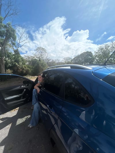 A young woman in a light blue shirt and jeans sits on the edge of the driver's seat of a blue hatchback parked on a dirt road. The woman is smiling and looking at the camera. The car's driver's side door is open. The sky is bright blue with white clouds. There are trees in the background. The scene is located in Santa Ana, Costa Rica at the coordinates latitude: 9.927, longitude: -84.181.