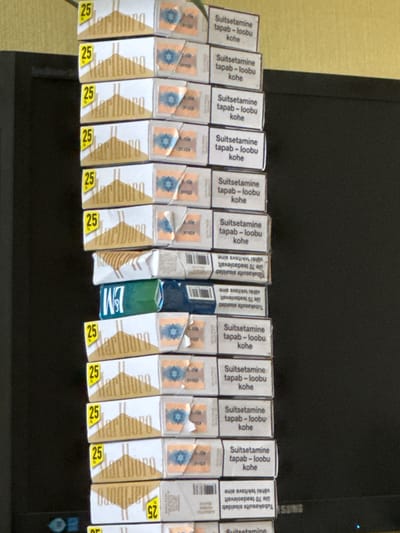 A stack of Marlboro cigarette packs is visible, with some packs showing "25" in yellow. The packs also have text in Estonian. The stack is positioned in front of a dark background, possibly a television screen. The overall scene appears to be indoors, with no specific city or location details discernible from the image itself. The lighting suggests an indoor environment.