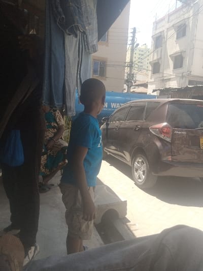 The image captures an urban street scene in Tudor ward, Kenya, under bright daylight. In the shaded foreground, several pairs of blue denim trousers hang from an overhead line or rack. To the far left, a person clad in dark attire stands, partially obscured, with a blue object visible by their leg. Behind this figure, another individual in a patterned brown, blue, and white dress is seated or crouching. A light-colored, possibly concrete, block with an arched opening rests on the paved ground.
Centrally, a young boy, appearing to be African, stands facing away from the camera, looking towards the right. He wears a blue t-shirt and light-colored cargo shorts. To his right, a brown Toyota Ractis station wagon is parked, oriented towards the left, with its rear passenger side visible. A partial yellow license plate, ending in "8", is on the tailgate, which also displays the "Ractis" model name. Behind the vehicle, a large blue cylindrical tank, likely a water storage unit, is visible, bearing the partially discernible text "KAN WA" and the number "188427029".
The background features multiple multi-story buildings, some with visible windows, and utility poles heavily laden with wires. The ground throughout the visible street area is paved with light-colored blocks. A blurred portion of a person's leg or lap is visible in the lower right foreground, suggesting the camera's perspective.