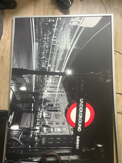 A framed black and white poster of the London Underground at Piccadilly Circus Station is leaning against a wooden floor. The poster depicts a nighttime scene with light trails from vehicles and illuminated buildings. The iconic London Underground logo is prominently displayed on the right side of the poster. The poster is oriented vertically. The image does not show any specific location in Gelsenkirchen, Germany.