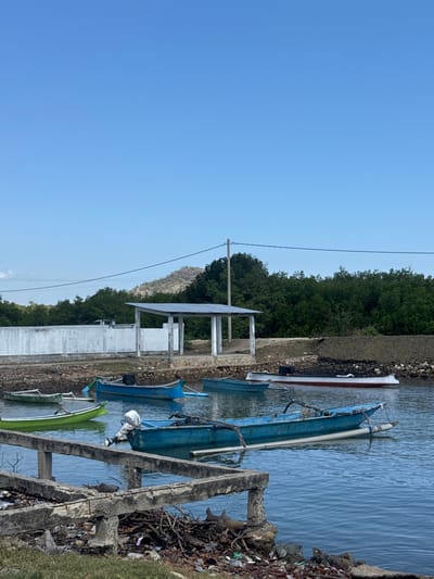 The image shows a body of water with several boats. In the background, there is a white wall, a covered structure with a metal roof supported by pillars, and lush green trees. Beyond the trees, a hilly landscape is visible under a clear blue sky. The foreground features a concrete structure with exposed rebar, debris, and dry vegetation. The water is calm and reflects the surrounding environment. There are no people visible in the image. The scene appears to be a coastal or riverine area in Kertasari, Indonesia, likely a small dock or mooring area.