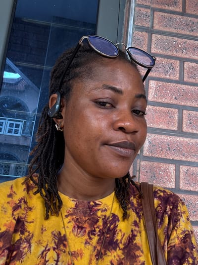 A close-up portrait shows a woman with dark skin and dreadlocks. She is wearing a yellow tie-dye t-shirt with a brown and purple pattern, a gold chain necklace, and a brown leather strap across her shoulder. She has sunglasses perched on top of her head and is wearing a black earbud. Her left ear has a silver hoop earring. She is positioned in front of a brick wall, and to her left, a blue reflective surface shows a building with white windows and architectural details, possibly a reflection. The scene appears to be outdoors. The woman's location in Ajara, Nigeria, is not determinable from the visual information.