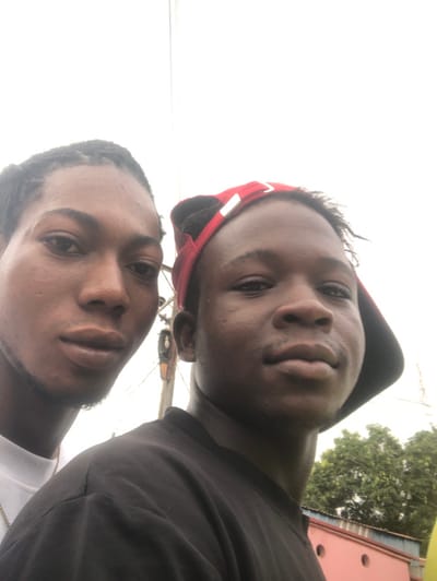 A close-up outdoor portrait depicts two young males in Oraukwu, Nigeria. The individual on the left has dark skin, dark braided hair, and wears a white shirt with a thin silver-colored chain, looking directly forward. The individual on the right, also with dark skin, wears a black t-shirt and a red baseball cap worn backward, looking towards the camera. The background features a bright, overcast sky, green tree foliage, a reddish-pink building with a white top edge, and visible utility poles with associated wiring, indicating an urban or semi-urban environment.