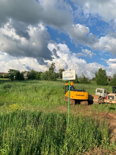 The image depicts a rural scene in the village of Svyatitsa, Russia, on a partly cloudy day. In the foreground, tall green grass dominates the landscape, with a few yellow wildflowers visible. A green signpost stands in the middle ground, bearing white lettering in Russian that reads "Соя мой источник" (Soy is my source) with an arrow pointing to the left.
Behind the signpost are several pieces of construction equipment. A yellow LiuGong excavator is positioned prominently, with its tracks resting on the muddy ground. To its right, a white truck with a raised cabin and a metal roller attachment is parked. Another piece of yellow construction equipment, possibly a bulldozer or another excavator, is partially visible to the far right.
In the background, a cluster of trees and several rustic houses suggest a residential area. The sky is filled with dramatic, dark grey clouds interspersed with patches of bright blue sky, indicating dynamic weather conditions. The overall impression is one of rural industry and construction work set against a natural, somewhat wild, backdrop. No people are visible in the frame.