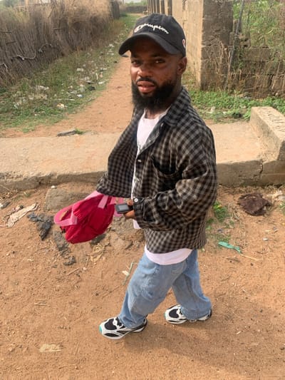 A male individual is observed walking on a reddish-brown dirt path in Eika-Adagu New Layout, Nigeria. He is an adult with dark skin and a full beard, looking directly towards the camera. He wears a black baseball cap with "Champion" embroidered in white, a white t-shirt, an unbuttoned grey and black plaid long-sleeved shirt, light blue jeans, and distinctive white, black, and light blue sneakers. In his left hand, he carries a red bag with pink straps. In his right hand, he holds a black feature phone. To the right of the path, a raised concrete curb or partial pathway is visible, with sparse vegetation and some scattered debris including plastic and fabric items. The background features more dry, reedy vegetation on the left, and unrendered concrete block wall structures or foundations on the right, suggesting undeveloped or partially constructed areas. The scene is illuminated by bright daylight.