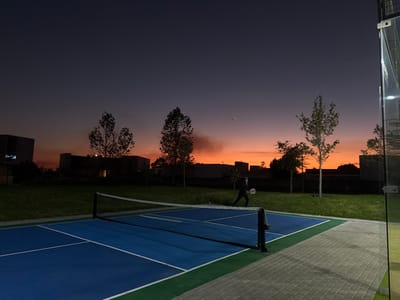 A person plays padel on a blue court with a net under a colorful sunset sky. Tall trees and buildings are silhouetted in the background. The court is surrounded by green grass and a tiled pathway. A glass wall is visible on the right side of the court. The sky displays hues of orange and dark blue.