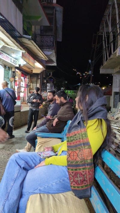 Here's a matter-of-fact description of the image:

**Location:** Dharamkot, India.

**Scene:** The image captures a nighttime street scene. A group of people are sitting on a blue wooden bench facing a street with shops. A woman with long dark hair, wearing a yellow sweater and a patterned shawl or scarf, is looking down. There are at least two men beside her, wearing jackets. One man is holding a phone. Further down the street, there are more people walking.

**Environment:** The environment consists of buildings, some illuminated with lights, indicating shops. The shops appear to be selling goods such as jewellery. The sky is dark, suggesting it is night. Overhead, there are electrical wires and building structures.