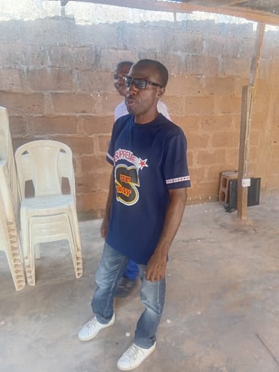 The scene depicts two adult males in a semi-enclosed, utilitarian area within Ayangba, Nigeria. In the foreground, an adult male with dark skin, short dark hair, and a slight beard, wearing black rectangular glasses, a dark blue short-sleeved t-shirt with "SUPREME IT'S MY WAY" graphics, blue jeans, and white sneakers, stands with his mouth slightly open, facing slightly to the viewer's right. His arms are down, hands loosely clasped. Behind and to the left of the primary subject, a second adult male with lighter skin tone is partially visible, appearing to smile. To the left of the foreground subject, a stack of four light-colored plastic chairs is present. The background features an unfinished light brown concrete block wall. The floor is a light gray concrete surface with scattered white debris. On the right side, a vertical wooden beam supports an overhead structure, with a black electrical outlet or box attached near its base. The area is illuminated by bright natural light.