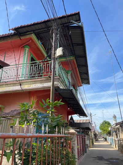 This image depicts a street scene in the city of Kota Kuala Simpang, Indonesia, under a clear blue sky with scattered white clouds. In the foreground, an orange metal fence with decorative finials runs horizontally across the frame, partially obscuring lush green foliage. To the left, a two-story building painted a vibrant coral pink with green accents dominates the view. It features a green metal railing on its balcony, supported by fluted green columns. A corrugated metal roof extends over the building's upper story. A utility pole densely covered with a tangle of black electrical wires stands prominently in front of the building. To the right of the building, a narrow paved street stretches into the distance, lined with houses of varying designs and colors. A lone figure on a motorcycle is visible further down the street. In the background, a minaret of a mosque rises above the rooftops, and mature green trees dot the landscape. The overall impression is one of a residential street in a tropical urban environment.