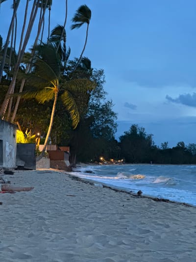 In the city of Tong Yang, Thailand, a sandy beach stretches along a coastline as dusk settles. The sky is a deep blue, transitioning to twilight, with wispy clouds visible. Palm trees, their fronds rustling in the wind, line the left side of the beach, some illuminated by warm yellow lights. To the left, a concrete structure, possibly a building or wall, bears some graffiti. Further back, there are more trees and some low wooden structures, with lights suggesting habitation or businesses. The ocean waves are active, crashing onto the shore, with foam and whitecaps visible. Scattered debris, like driftwood and a coconut, lie on the sand near the water's edge. In the foreground, a person's legs and feet are visible, lying on the sand, indicating they are relaxing on the beach. Their body is mostly out of frame, but they appear to be wearing a red garment. The overall scene conveys a serene, tropical evening atmosphere.