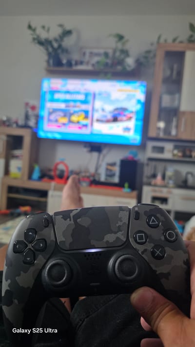 The media file shows a person holding a PlayStation 5 controller in a room. The controller has a camouflage pattern. In the background, a television screen displays what appears to be a video game menu with colorful graphics. The room has shelves with various items, including plants. The lighting suggests it could be daytime or evening. The setting is indoors, likely a living room or entertainment area. The focus is on the controller and the game being played, implying an activity of playing video games. Visible text on the media file reads "Galaxy S25 Ultra" on the right side, which is likely a watermark from the device used to capture the image.