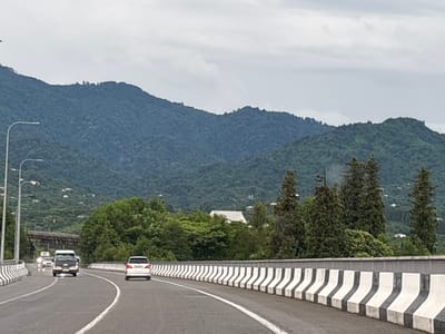 A wide-angle shot captures a view from a highway in Chakvi, Georgia, on a cloudy day. In the foreground, the road curves to the right, bordered by a concrete barrier with alternating black and white stripes. Several vehicles are visible on the highway, including a white SUV in the right lane, a black and white van in the left lane, and two other white vehicles further ahead.
The middle ground features a lush green forest that climbs up the slopes of rolling mountains in the background. Some scattered white buildings are visible on the mountainsides, suggesting a rural or suburban setting. Tall, slender trees are interspersed with denser foliage.
Overhead, the sky is overcast with thick, gray clouds, indicating a possibly cool or rainy day. The overall mood is serene and somewhat muted due to the weather conditions. The scene depicts a typical travel moment on a highway winding through a mountainous, verdant landscape in Georgia. There is no visible text in the image.