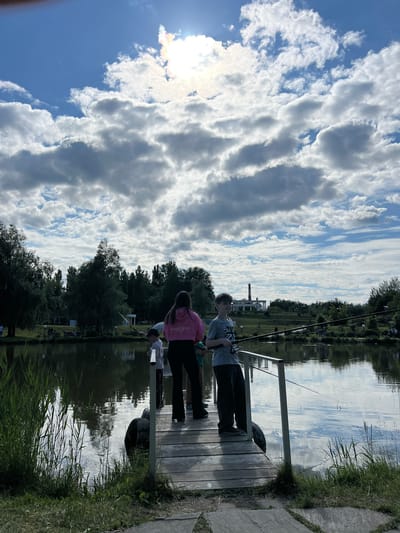 The image depicts an outdoor recreational scene on a partly cloudy day in Cheboksary, Russia. The foreground features a wooden dock extending into a calm body of water, likely a pond or lake, reflecting the sky and clouds. Tall green reeds and grass grow along the immediate shoreline.
On the dock, three individuals are present. A young boy, wearing a grey t-shirt with a Union Jack design, dark trousers, and glasses, stands to the left, holding a fishing rod. He appears to be focused on the water. Behind and slightly to his right stands a taller person, likely an adult female, wearing a pink hooded sweatshirt with "GYMAESTHETICS" printed on the back and dark trousers. Her back is mostly to the camera. Between these two, a smaller child in a light-colored t-shirt and a dark cap is partially visible, also looking towards the water. A fishing line from one of the rods extends into the water, creating small ripples. The dock's railing is metallic and white. Two large, dark, circular objects, possibly tires, are partially submerged or positioned next to the dock's support structure.
The midground consists of the water body surrounded by dense green foliage, primarily trees. On the far bank, several indistinct figures suggest other people are present. In the background, beyond the treeline, distinctive urban structures are visible. To the left, a prominent white building with classical architectural elements, including columns and a domed or complex roof, stands amidst greenery. To its right, a tall industrial chimney with red and white horizontal stripes rises above the landscape. The sky above is blue with significant white and grey cumulus clouds. The sun is visible in the upper right quadrant, partially obscured by clouds, causing a bright glare and slight lens flare with a visible rainbow diffraction effect.