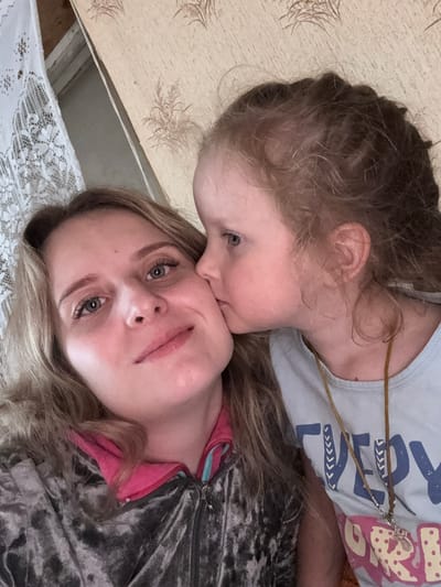 A close-up, overhead, indoor selfie shows two females, an adult and a child. The adult, positioned on the left and slightly out of focus, has blonde hair and is looking at the camera. She is wearing a gray velvet jacket with a pink collar. The child, on the right and in focus, is leaning in to kiss the adult's cheek. The child has blonde hair and is wearing a light blue t-shirt with blue and pink lettering. The background features textured wallpaper and a white lace curtain. The image appears to be taken in a domestic setting, likely in Severnyy, Russia.