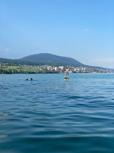 In a serene lake setting, a person is paddleboarding on a calm blue body of water. The paddleboarder, wearing swim trunks and a hat, stands on a yellow board, holding a paddle. In the distance, a town with white buildings nestled against a lush green hillside can be seen. Two individuals are swimming in the water further from the shore. The sky above is a clear, bright blue, suggesting a sunny day. The overall scene evokes a sense of leisure and outdoor recreation on a pleasant day, likely in the summer. The location context is Milvignes, Switzerland.