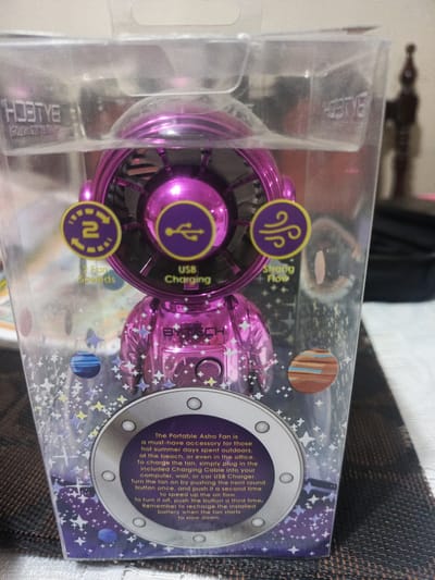 The image shows a close-up, slightly angled view of a pink, astronaut-shaped portable fan packaged in clear plastic. The fan is the central subject, with its metallic pink body and silver helmet clearly visible. It is adorned with various icons on its front, including one indicating "2 Fan Speeds" and another for "USB Charging," and a third symbol representing airflow, labeled "Strong Flow." The packaging is decorated with a starry, cosmic design featuring planets. The product is placed on a textured, dark brown surface, possibly a table or placemat. In the background, the lighting suggests an indoor setting, with a blurry, dark object to the right that could be furniture or an appliance. The text "BYTECH" is visible on the packaging and on the fan itself. The overall impression is that of a product display, likely in a retail environment or home. There is no indication of the location within Guayaquil, Ecuador.