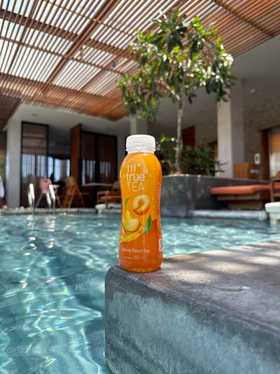 A bottle of "TH true TEA Natural Peach Tea" is placed on the edge of a swimming pool. The bottle is orange and has a label with a picture of peaches. The swimming pool water is clear and blue, with sunlight reflecting on the surface. In the background, there is a wooden pergola, trees, and lounge chairs. The scene appears to be set in a resort or hotel in South Quy Nhon Ward, Vietnam, judging by the architectural style and vegetation. There are no people clearly visible in the immediate vicinity of the bottle, though some figures are blurred in the background near the pool and building structures. The lighting suggests it is daytime, likely with bright sunlight.