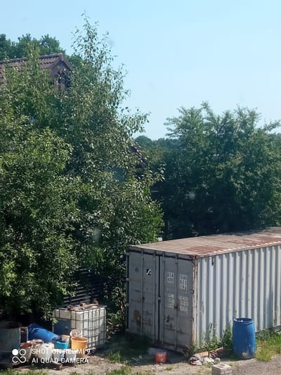 The image shows an outdoor scene with a shipping container, a large plastic water tank, several barrels and buckets, and lush green trees. A roof of a building is visible through the trees. The scene appears to be in a rural or industrial setting. The sky is clear and blue, suggesting it is daytime and the weather is fair. The shipping container has markings on it, including "USA 4310" and "SEAU 823 796 15". There is also a watermark in the lower left corner that reads "SHOT ON REDMI 9 AI QUAD CAMERA". The overall impression is one of a storage or work area, possibly on a farm or in a yard. No people are visible in the frame.