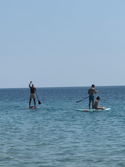 The image depicts two individuals paddleboarding on a body of water, likely the sea, under a clear sky. The scene is oriented such that the water fills the right side of the frame and the sky the left, suggesting a strong vertical orientation to the original capture.
In the foreground, a person is standing on a paddleboard, facing away from the camera and towards the water. They are holding a paddle, with the blade submerged, indicating they are actively paddling. This individual is wearing a white cap and light-colored shorts.
Further in the distance, on another paddleboard, a man and a woman are present. The man is in a seated position, holding a paddle, while the woman is lying down on the board. They are both looking towards the water. The man appears to be wearing shorts.
The water exhibits ripples and texture, suggesting a gentle movement. The sky is a consistent, pale blue, with no visible clouds. The lighting suggests it is daytime, likely late morning or early afternoon, given the bright illumination. The location context places this scene in Mirište, Montenegro, which is a coastal area. No text is visible in the image.