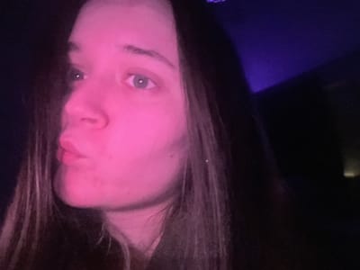 A female subject, appearing young, is depicted from the upper torso upwards, with her head turned to the viewer's left, and her gaze directed upwards and to the left. Her lips are pursed. The right side of her face (viewer's left) is intensely illuminated by a pink light source, casting a magenta hue across her skin. Her right eye (viewer's right) is visible, featuring a light-colored iris and a distinct pupil. Long, dark hair frames her face and extends downwards. The background is predominantly dark, with a distinct area of purple light visible in the upper right portion of the frame. The scene is located in the city of Pushchino, Russia.