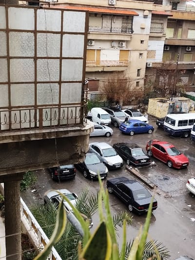 The image depicts an elevated view of an outdoor parking area within the city of As-Salihiya Municipality, Syria. The scene appears wet, suggesting recent rainfall.
In the immediate foreground, from a balcony perspective, a large multi-paned structure with frosted or textured glass and a dark frame partially obscures the upper left view. A thin, dark cable or wire runs vertically down its center. Below this, a concrete balcony edge exhibits chipped and deteriorated sections, with a decorative dark metal railing visible. Green foliage, likely Sansevieria and palm fronds, is present at the bottom of the frame, originating from below the balcony.
The midground is dominated by a wet, asphalt or concrete parking lot containing numerous parked vehicles. These include a white utility van, a silver SUV, a white sedan, a blue sedan, a black sedan, a red sedan, a dark blue and white mini-bus, and several other black and silver sedans and hatchbacks. One individual is observed standing near the rear of the blue sedan, appearing to interact with the vehicle. Another figure is faintly visible near the silver SUV. The ground throughout the parking area is visibly wet with scattered puddles. A broken concrete curb section lies on the ground near the center-right. A yellow corrugated metal container or skip is positioned at the far right of the parking area.
In the background, multi-story residential buildings rise, featuring balconies with metal railings, various windows (some with closed roller shutters or blinds), and multiple external air conditioning units. Sparse, bare-branched trees or large shrubs are situated behind the parking lot. The sky overhead is overcast, providing diffuse lighting to the scene.