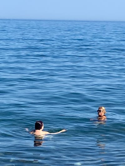 Two individuals are submerged in the calm blue waters of the Mediterranean Sea, with the faint horizon of Marbella, Spain, visible in the distance. The person on the left, with dark hair, is facing away from the camera and appears to be swimming. The person on the right, with blonde hair and sunglasses, is facing towards the camera, their upper body visible above the water. Both individuals are wearing swimwear. The water is clear with gentle ripples on the surface, reflecting the clear sky above.