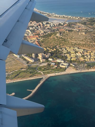 An aerial view captures the coastal city of Cagliari, Italy, with an airplane wing prominently in the foreground, obscuring part of the scene. The image displays a dense urban area with numerous buildings, predominantly light-colored and flat-roofed, clustered together on a hillside. The city appears to be built along a bay or harbor, with the turquoise waters of the sea visible in the lower portion of the frame. A long, curved breakwater made of rocks extends into the water, protecting a portion of the coastline. Beyond the immediate urban sprawl, salt pans are visible, characterized by rectangular, shallow bodies of water. The landscape is a mix of natural vegetation, including trees and shrubs, interspersed with the built environment. Boats can be seen anchored in the distance, suggesting maritime activity. The overall impression is of a sunny day with clear skies, casting shadows that highlight the textures of the buildings and landscape.
