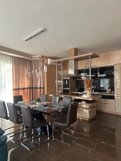 The image depicts a modern, open-plan dining and kitchen area in an apartment located in Brca, Montenegro. A large, rectangular dining table with a glass top and a dark base is positioned in the foreground, surrounded by eight dark brown leather chairs with chrome frames. The table is set with dark placemats and cutlery. A small green potted plant and a woven basket are placed in the center of the table.
Behind the dining area, a sleek, contemporary kitchen features high-gloss cabinetry in cream and black. A prominent stainless steel range hood is installed above the stovetop on a central island that also incorporates shelving and storage. Various kitchen appliances, including an oven and microwave, are visible. Decorative dried wheat stalks in a woven vase sit on the island. To the right of the kitchen, a glass-fronted cabinet displays glassware and bottles of alcohol, suggesting a bar area.
The room is illuminated by natural light streaming through sheer white curtains and brown-toned drapes on the left side, and by a linear pendant light fixture above the dining table. The flooring is dark, glossy tile, reflecting the light and the furnishings. The walls are textured in a warm, sandy beige and a darker, almost coppery hue. The overall impression is one of contemporary luxury and spaciousness.