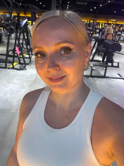 The image shows a woman taking a selfie in a gym. She is wearing a white tank top and has blonde hair. She is wearing makeup and earrings. The gym appears to be well-lit and equipped with various exercise machines. In the background, other gym-goers can be seen using equipment. The overall environment suggests a modern fitness facility. The city is Komsomolsk-on-Amur, Russia.