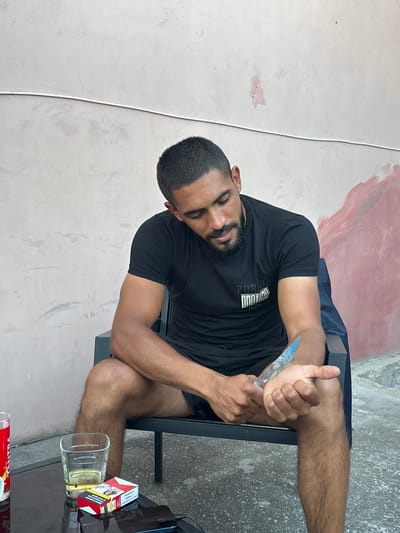 A man with dark hair and a beard sits on a chair in front of a textured wall. He is wearing a black t-shirt and black shorts. In his right hand, he holds a knife, which he appears to be using to cut his left forearm. On a glass table in front of him are a glass of amber liquid, a pack of cigarettes, and a mobile phone. The environment appears to be an outdoor seating area, possibly a patio. The wall behind him is painted a light color, with some pinkish-red patches. The ground is concrete.