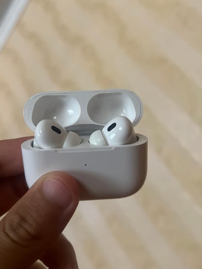 In an indoor setting, a person's hand is holding an open white charging case for wireless earbuds. The case is white and slightly rounded. Inside the case, two white wireless earbuds are visible, nestled in their charging slots. One earbud is more fully visible, showing its white body and a small black vent or sensor. The other earbud is partially obscured by the case. The background is a blurred, light brown textured surface, possibly a wall or furniture, with subtle variations in tone. The lighting suggests an indoor environment, potentially with natural light contributing to the overall brightness. The image is a close-up, focusing on the earbuds and the hand holding the case. No text is visible in the image.