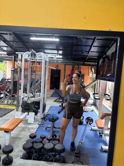 A woman is taking a mirror selfie in a gym located in Kampot, Cambodia. She is wearing a grey and black striped sports bra and brown athletic shorts. She has headphones on and is holding her phone in her right hand, with her left hand on her hip. The gym appears to be open-air with a corrugated metal roof supported by metal beams. Equipment visible includes a weight machine, dumbbells, a weight bench, and a barbell. The walls are painted a bright orange. A ceiling fan is visible in the background. There is a television screen mounted on the wall displaying an image. The floor is covered with blue and yellow mats. Outside, there are trees and foliage visible.