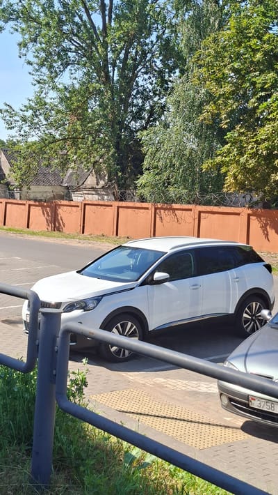 A white Peugeot 3008 SUV is parked in a paved lot in front of a coral-colored wall topped with barbed wire. The car is positioned in the foreground, angled slightly to the right. Behind the car, a row of trees with green foliage creates a natural backdrop. In the distance, buildings with tiled roofs are visible. The sun is shining, casting shadows on the ground. The license plate of the car is partially visible, with the letters "BY" indicating Belarus. The scene appears to be in the city of Smalyavichy, Belarus.