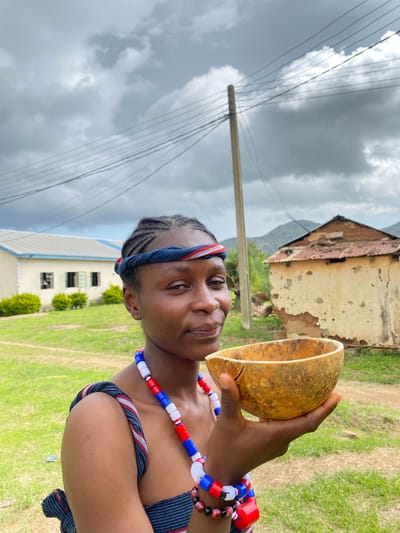 A person is holding a gourd in their right hand, tilted as if to drink from it. They are wearing a beaded necklace of red, white, and blue beads, and a blue and white patterned garment. A matching headband with red and white stripes is worn across their forehead. The person's braided hair is visible. The background shows a rural scene with a dilapidated building with a rusty roof and peeling paint. In the middle distance, a light-colored building with a few windows is visible, surrounded by green grass. A wooden pole with multiple wires stretches across the cloudy sky, indicating the presence of electricity. The sky is overcast, suggesting a potentially rainy or stormy day. The person is looking towards the camera with a slight smile, conveying a sense of calm or anticipation. The overall setting suggests a village in Wang, Nigeria.