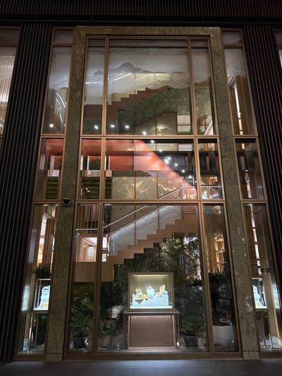 The image shows a multi-story building facade in the Jinjiang District of China. The facade is composed of large, rectangular glass panels, framed by a material that appears to be bronze or a similar metal. The frames are inlaid in what looks to be green marble or a similar stone material. Inside, stairs with metal railings are visible through the glass. Additionally, there are displayed items and plants. The interior design suggests the building is either a high-end retail location or an upscale residential structure. The image appears to be taken at night, and there are reflections in the glass, suggesting the interior is well-lit.