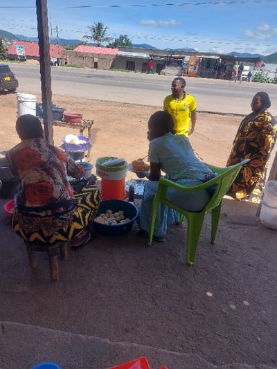 The scene depicts a roadside food stall in Kimamba, Tanzania, under a clear to partly cloudy sky. In the immediate foreground, a person is seated on a small wooden stool with their back to the viewer, wearing a patterned garment with orange and black motifs. This individual is positioned behind a small wooden table bearing various cooking implements and containers, including metal pots, plastic bowls (one purple bowl containing a yellow substance, another blue bowl holding multiple white spherical food items), and a white bucket.
To the right, a second person is seated in a light green plastic chair, facing left and partially away. This individual wears light blue clothing and holds a plate with food in one hand and a small bottle in the other. Adjacent to the seated individual and closer to the road, a male stands in a bright yellow t-shirt with a graphic design, facing left. To the right of the standing male, a female stands, wearing a dark patterned fabric with prominent yellow/gold floral designs, also facing left.
Behind the immediate activity, an asphalt road runs horizontally across the frame. A dirt shoulder separates the food stall area from the road. Across the road, a line of single-story buildings with various roof types (some red, some flat) is visible. A dark-colored vehicle is partially visible on the far left side of the road. In the distance, hills and mountains are silhouetted against the horizon. Vegetation, including a palm tree, is scattered throughout the background. Shadows indicate bright overhead sunlight.