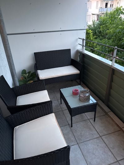 The image depicts a balcony in Höhenkirchen-Siegertsbrunn, Germany, furnished with a dark rattan sofa and two armchairs, each with white cushions. A glass-top coffee table with a woven rattan base sits in front of the sofa. On the table are a pink ashtray and a decorative glass jar with a patterned surface. A potted plant with green leaves is visible behind the sofa. The balcony has a tiled floor and a metal railing. In the background, trees and a multi-story building with balconies are visible. The overall scene suggests an outdoor seating area on a residential balcony.
