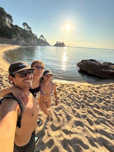 This is a wide-angle, eye-level selfie taken on a beach in Castell d'Aro, Platja d'Aro i s'Agaró, Spain. Three people are visible. In the foreground on the left is a man wearing a dark baseball cap with the word "Supreme" on it, dark sunglasses, and a silver chain necklace. He is shirtless and appears to be wearing dark shorts. His left arm is around the shoulders of a woman standing to his right. She is wearing sunglasses and a black tank top, and a gold chain necklace. To the right of her and slightly behind her is a third person, appearing to be a woman, wearing a baseball cap and a purple and white striped bikini. All three individuals are smiling and looking towards the camera.
The background shows a sandy beach with visible footprints and ripples from the tide. The sea is calm and reflects the bright sun, which is high in the sky and creating a lens flare. A rocky outcrop with trees on top is visible in the mid-ground to the right, and a small island with trees is in the distance. The shoreline curves to the left, with a vegetated cliff face dotted with buildings and trees rising on the far left. The lighting suggests it is either early morning or late afternoon.