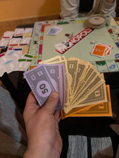 The image depicts a Monopoly board game in progress, located in the city of Gerakas Municipal Unit, Greece. A first-person perspective shows a human hand in the foreground holding a fanned stack of Monopoly currency, consisting of purple 50-denomination bills on the left, numerous tan 100-denomination bills in the center, and orange 500-denomination bills visible at the bottom right. The Monopoly game board is laid out on a light-colored wooden table. Various game components are visible on the board: property deeds are organized in multiple stacks on the left side, chance cards (orange with a question mark) and community chest cards (blue) are stacked in the center-right. Two white dice, green plastic houses, and game tokens are scattered on the board. The prominent red "MONOPOLY" logo is visible across the center. In the blurred background, another individual wearing a light-colored long-sleeved garment is partially visible, seated at the table, near a small, round, lidded woven container. The immediate environment includes a section of a light brown wall or floor in the background and textiles consistent with clothing on the person holding the money (grey on the left, dark with white stripes on the right).