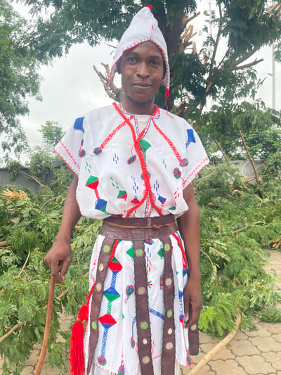 A man stands outdoors, wearing a white tunic with red, blue, and green embroidery, a leather belt, and a white conical hat. He holds a wooden stick in his left hand. The background consists of trees and branches, with a paved ground and a wall visible. The man appears to be in a yard or open space with fallen vegetation, possibly indicating recent storm damage or landscaping. The setting is likely in Takatuku Madorawa, Nigeria.