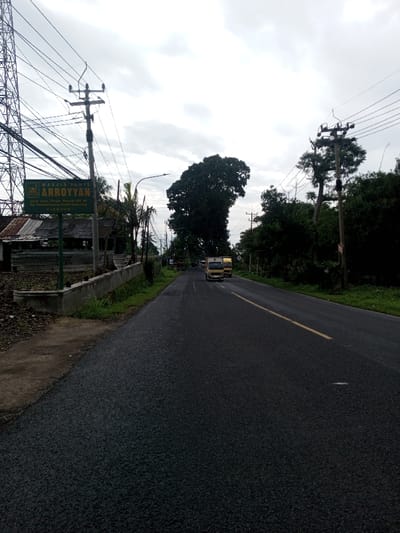 The scene depicts a straight asphalt road under an overcast sky. The road surface appears wet and features a single solid yellow line on its right side, defining a single lane of travel in the visible direction. In the distant center, two light-colored trucks are observed in motion, traveling away from the viewer.
On the left side of the road, a green rectangular sign is prominently displayed, reading "MASJID JAMI ARROYYAN" at the top. Below this, smaller text specifies the address: "Dusun Ciwiru, Parereja, RT.04 RW.01 Ds. Kertamulya, Kec. Lakbok CIAMIS". This indicates the location is Ciamis, Indonesia, which contradicts the requested location of Kamji Na-Yekha, Bhutan. Behind the sign and to its left, a structure with a corrugated metal roof and weathered wooden walls is partially visible. A tall metal lattice tower stands on the far left, connected to multiple utility poles by numerous overhead wires that traverse the scene. A low concrete barrier or retaining wall separates the road from the ground on the left, which consists of dirt and sparse vegetation.
The right side of the road is bordered by a dense line of green foliage and various trees, some of which are tall. Several utility poles with overhead wires are also present along this side. The sky above is uniformly gray with heavy cloud cover. No individuals are visible in the image.