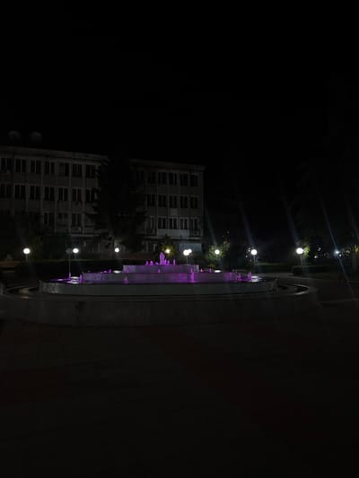 The image depicts a nighttime scene in Elhovo, Bulgaria, featuring a building, a fountain, and street lighting. The building is multi-storied with a grid-like pattern of windows, and an air conditioning unit mounted on the walls. The fountain is illuminated with purple lights, with water visibly spouting. A line of spherical streetlights runs parallel to the fountain and the building. The surrounding environment is dark.