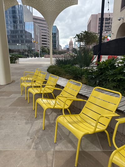 A row of yellow metal chairs lines a stone walkway. Lush greenery sits behind the chairs. In the background, city buildings and a state capitol building are visible under a cloudy sky. A white, perforated structure stands to the left. The overall impression is an urban outdoor space designed for relaxation and contemplation.