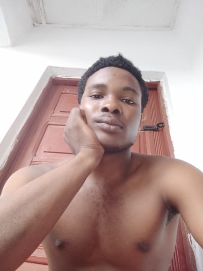 This is a medium-shot, forward-facing selfie of a young Black man with short, curly hair and dark eyes. He is shirtless and appears to be indoors, leaning his right arm on his chin with his hand supporting his jaw. The background consists of a reddish-brown wooden door with vertical panels and a black latch. The lighting suggests it is daytime, and the overall mood is relaxed and contemplative. The setting appears to be a home in Orodo, Nigeria, given the provided location context. The man's skin is smooth and appears healthy, with a slight sheen that might indicate perspiration or natural oil. The focus is primarily on the man's face and upper body, with the background slightly out of focus. There is no visible text in the image.