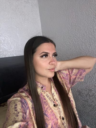 A woman with long, dark hair and dramatic false eyelashes poses with her right arm behind her head. She is wearing a button-up shirt with a psychedelic pattern of purple, pink, and yellow. The background is a textured gray wall. The image appears to be taken indoors. There is no indication of the city of Chalda, Russia, in the image.