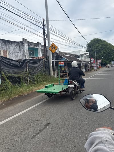 A motorcyclist wearing a white helmet and a black jacket rides a motorcycle with a green trailer attached. The motorcyclist is in the foreground of the image, moving from left to right on a paved road. The road has a white line marking the lane division. In the background, there are buildings, trees, and power lines. A yellow diamond-shaped sign with two figures on it, indicating a pedestrian crossing, is visible on the right side of the road. The sky is overcast. The reflection of the motorcyclist's face in the mirror shows them wearing a mask. The scene appears to be on a street in Ajuen, Indonesia.