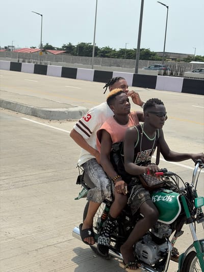 On a clear, sunny day in Jakande, Nigeria, three males are depicted riding a green and black motorcycle on a paved, multi-lane road.
The driver, a dark-skinned male with short dark hair, wears black wraparound sunglasses, a black basketball jersey with "BULLS" text, dark shorts (with "BALENCIAGA" visible), black patterned socks, and black flip-flops. A brown, designer-style crossbody bag is slung over his right shoulder. He grips the handlebars of the motorcycle.
Seated directly behind the driver is the middle passenger, a lighter-skinned male with short bleached blonde hair. He wears a light orange sleeveless top, dark shorts, and a yellow beaded bracelet on his left wrist. He is looking towards the left of the frame, with his right hand raised near his temple.
The rear passenger, a lighter-skinned male with dark dreadlocks, is seated behind the middle passenger. He wears a white short-sleeved t-shirt with "23" on the sleeve, grey patterned shorts, and black slide sandals.
The motorcycle itself is green and black, displaying "K-TOK" on its fuel tank and a visible Bajaj engine.
The environment consists of a wide, light grey asphalt road with clear white lane markings. A concrete road barrier, painted in alternating black and white sections, separates the road from the background. Several tall, dark streetlights with modern fixtures line the road. In the mid-ground, grey block walls, green foliage, and structures with reddish-brown rooftops are visible. Distant grey vehicles are partially discernable on another section of the road in the far background.