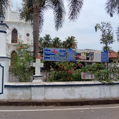 The image shows the exterior of St. Anthony's Church in Siolim, Marna, India. The white church is partially obscured by palm trees and other vegetation. A white wall with a blue-trimmed top runs along the front of the property, with a white cross placed atop it. Behind the wall, signs display church mass timings and architectural details. A "No Parking" sign is visible amidst the plants and iron fence behind the wall. The foreground features a paved road with a white line marking its edge.